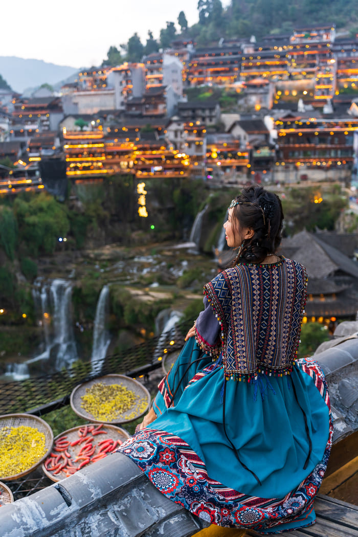 Una mujer vestida con ropa tradicional china sentada de espaldas mirando hacia la antigua ciudad iluminada de Lijiang y sus cascadas al atardecer.
