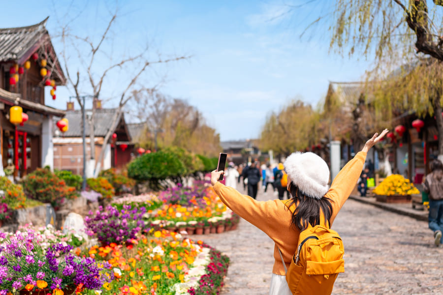 Una joven turista de espaldas, con un sombrero de fieltro blanco y una mochila naranja, se hace un selfie con los brazos extendidos en una calle peatonal del casco antiguo de Lijiang, China. La calle está llena de flores de colores y edificios tradicionales chinos de madera con tejados de tejas, bajo un cielo azul claro.