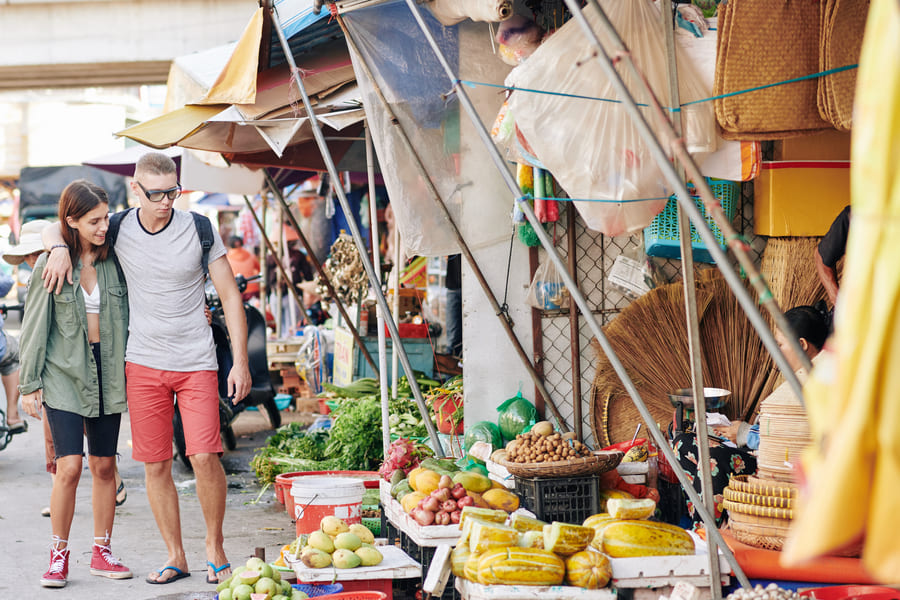 Pareja de turistas paseando por un mercado local al aire libre en China y observando puestos de frutas tropicales frescas.