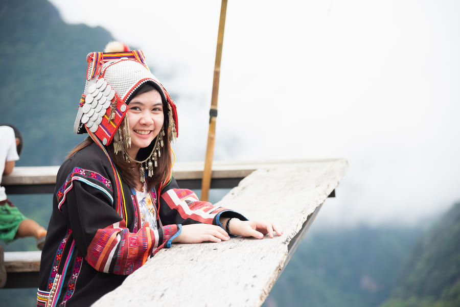 Mujer con vestimenta tradicional de una etnia de Yunnan sonriendo en un mirador con un paisaje montañoso al fondo.